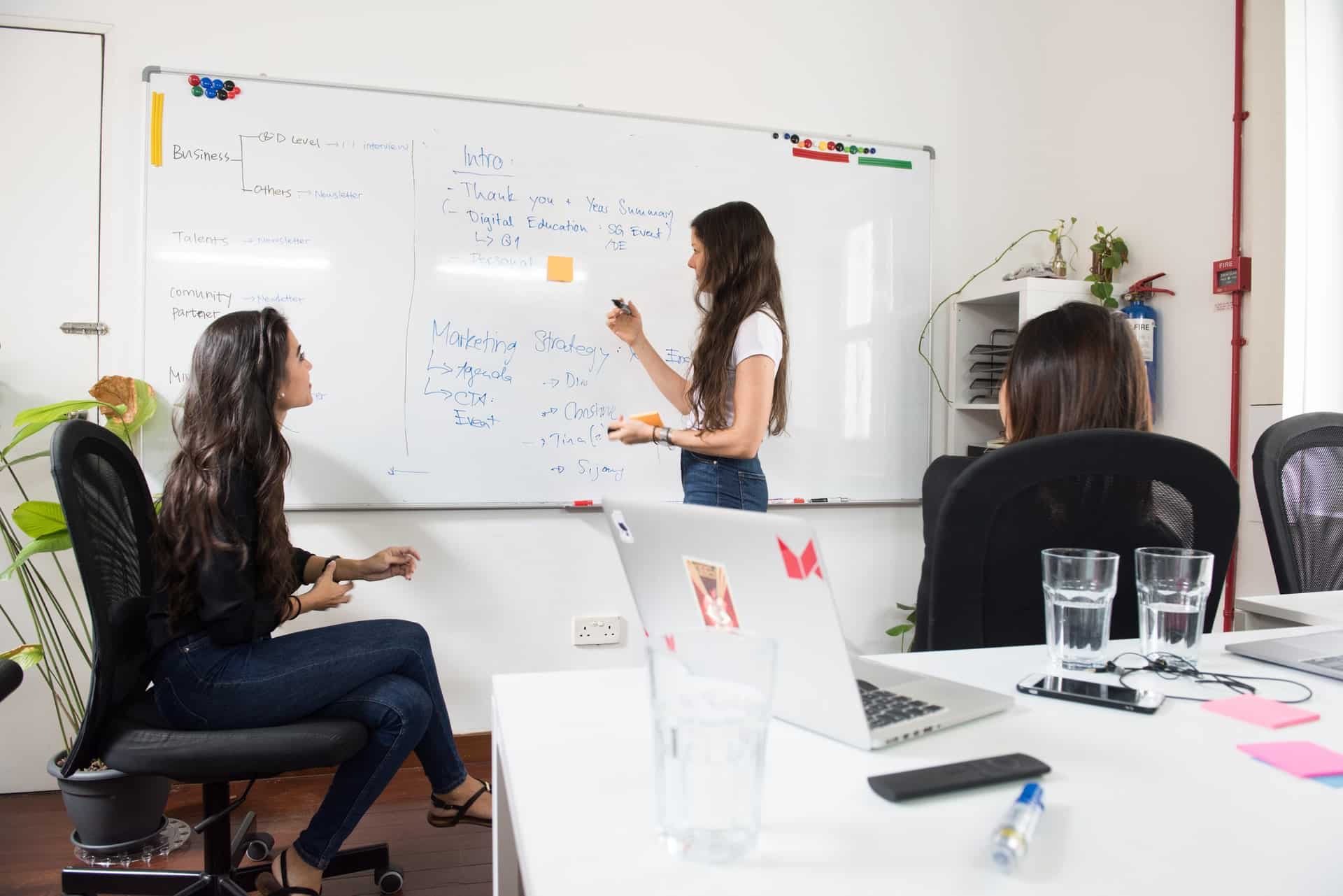 Image shows a woman writing on a whiteboard talking to a woman sitting down in an office chair, illustrating brainstorming.