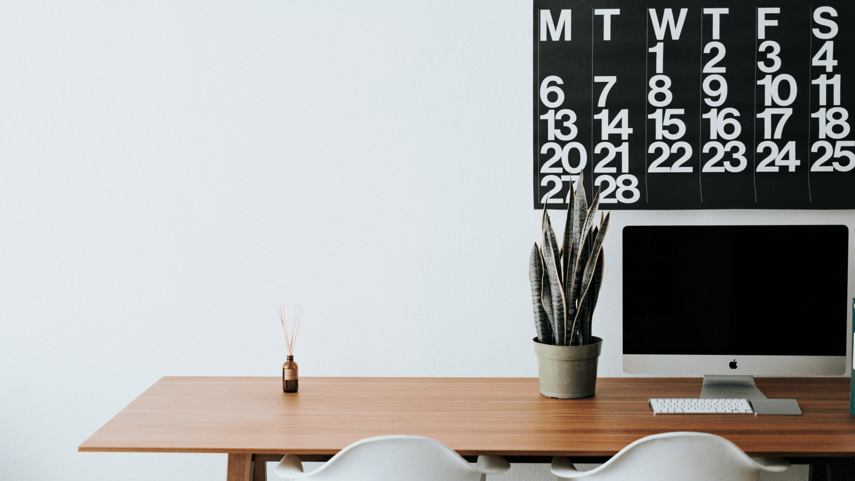 Das Bild zeigt einen zwei weiße Stühle vor einem braunen Schreibtisch auf dem eine Pflanze sowie ein ausgeschalteter Monitor steht. Dahinter ist ein Kalender an der Wand zu sehen.