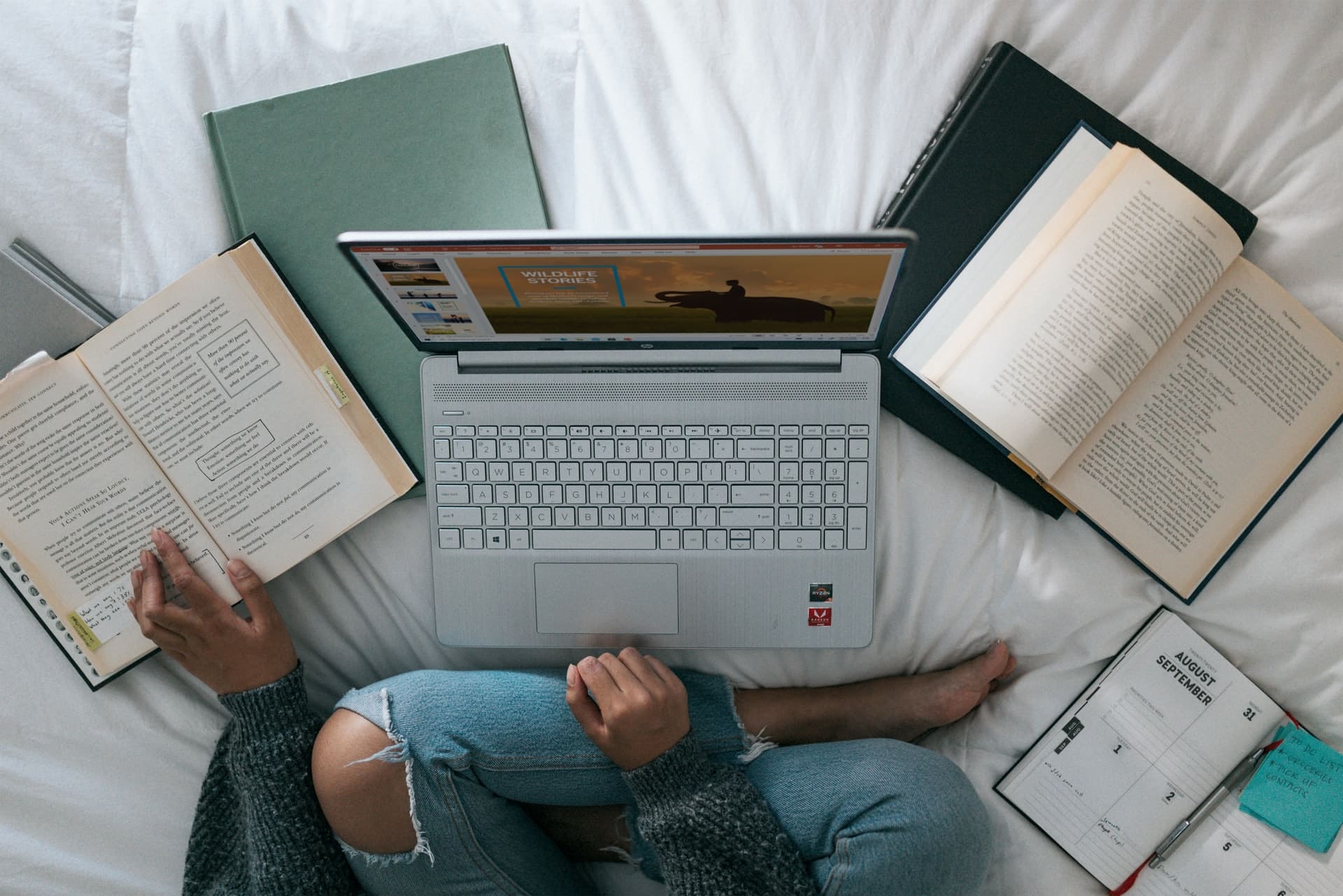 Das Bild zeigt eine Person vor ihrem Laptop. Neben dem Laptop liegen mehrere geöffnete Bücher.
