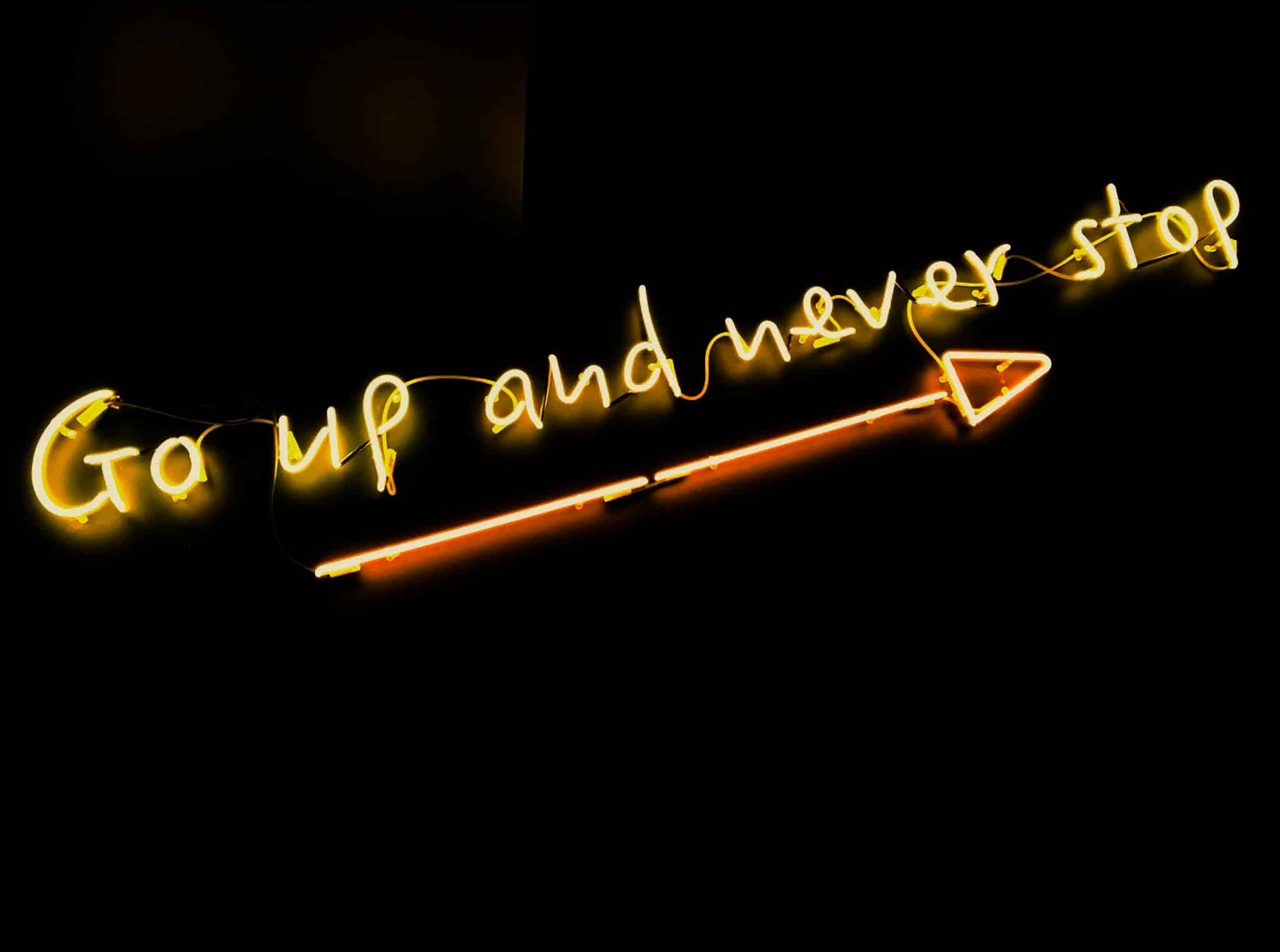 Das Bild zeigt den Satz "Go up and never stop", der erhellt in der Dunkelheit leuchtet. Darunter leuchtet ein Pfeil, der nach rechts oben zeigt und den Satz unterstreicht.