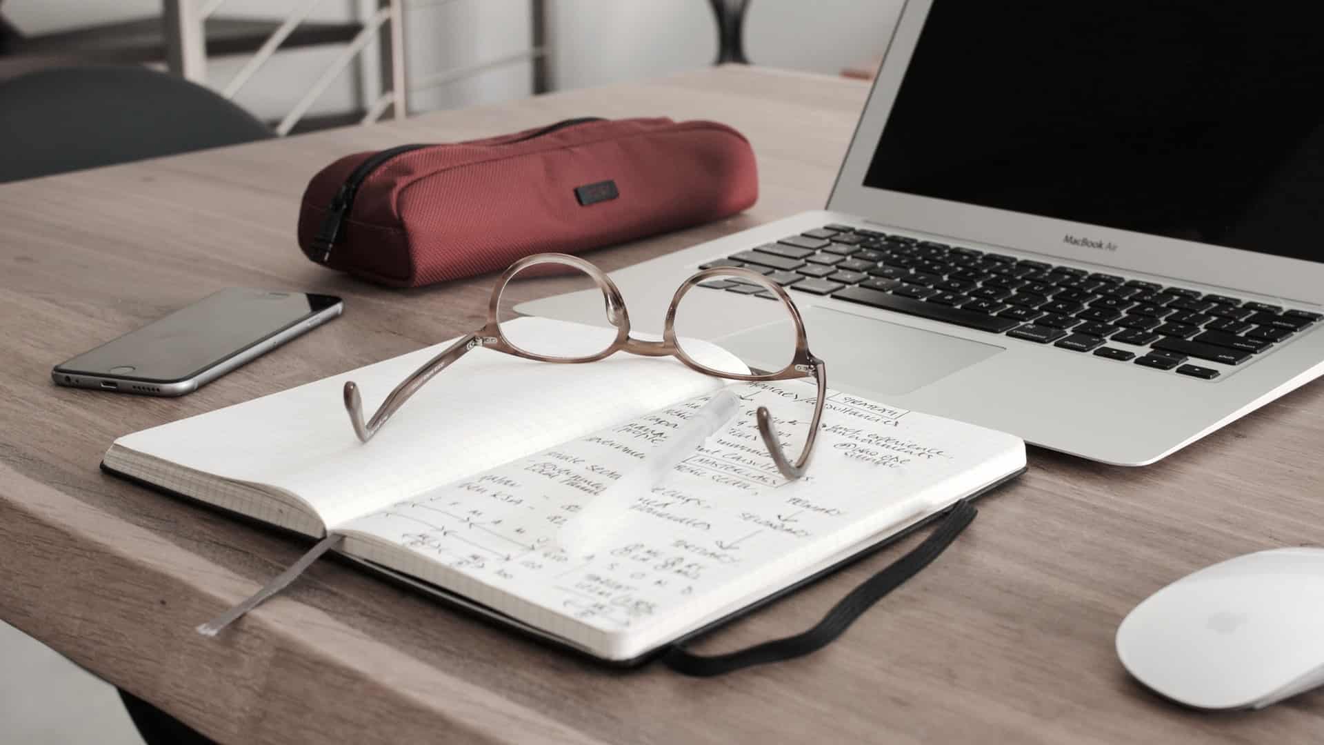 Image shows glasses on a notebook and an open laptop, illustrating the concept of academia.
