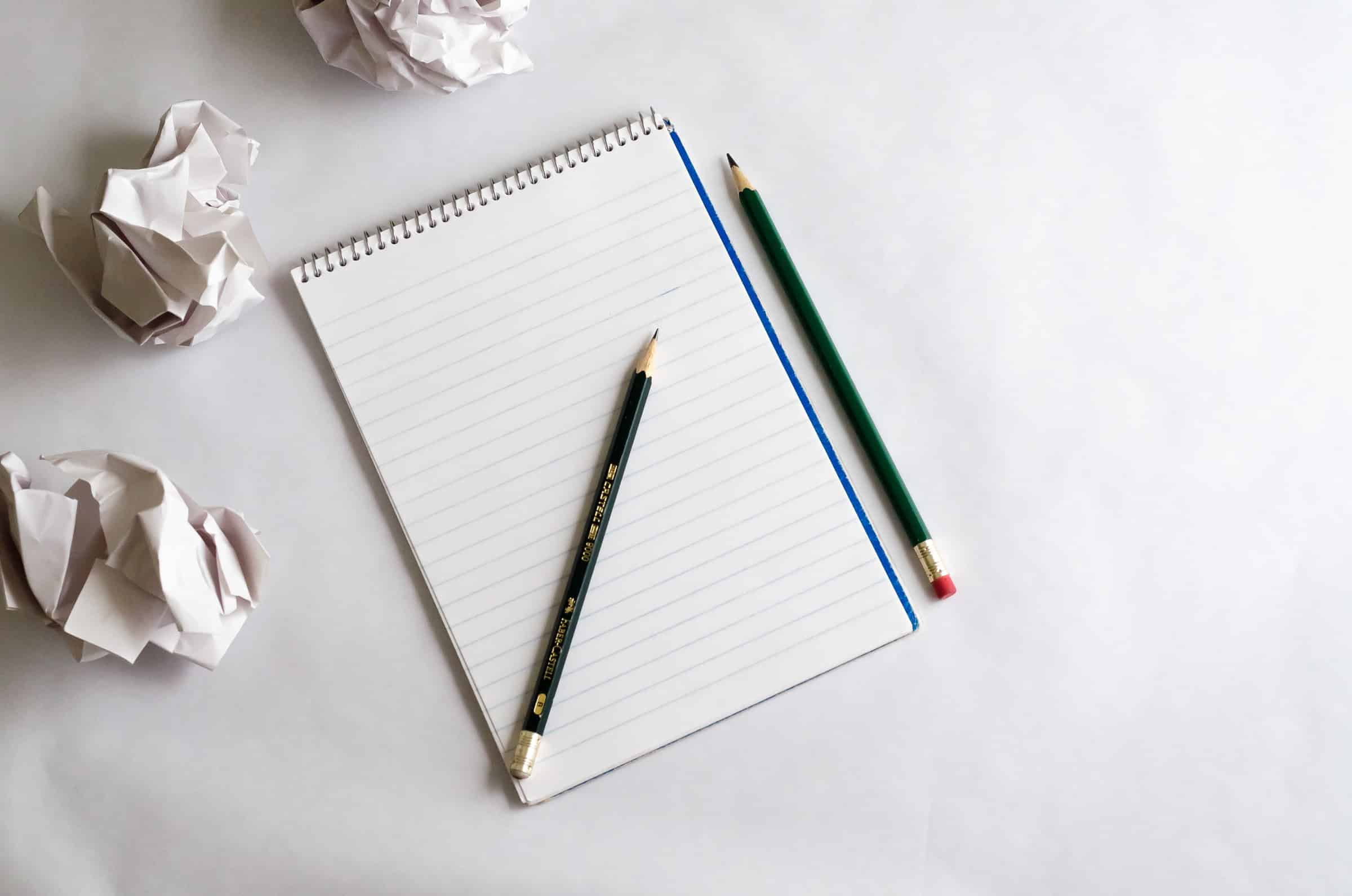 The image shows an empty notepad. Two pencils and three balls of crumpled paper are strewn around it.