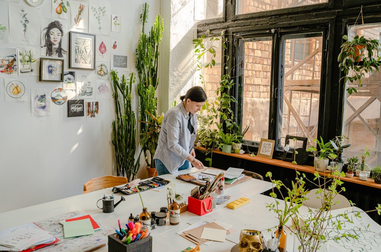 The image shows a woman working in a bright and lovely studio surrounded by plants and tools, giving us a picture of how a business owners life looks like.
