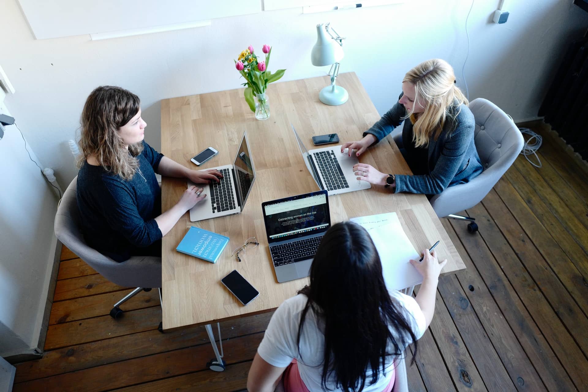 The image shows three women talking and working on their laptops.
