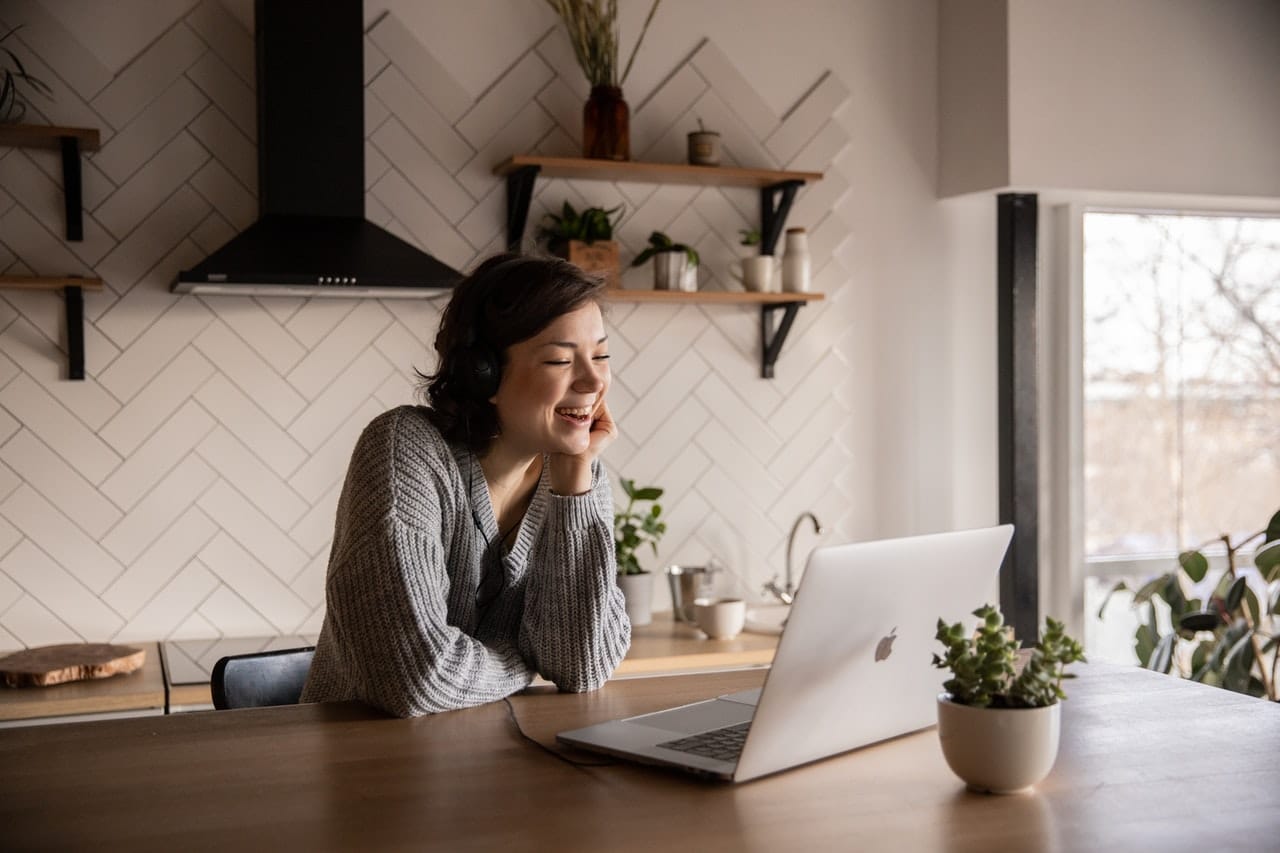 A woman is sitting in her minimal kitchen enjoying the online meeting with a big smile. It shows how virtual team-building games are important.